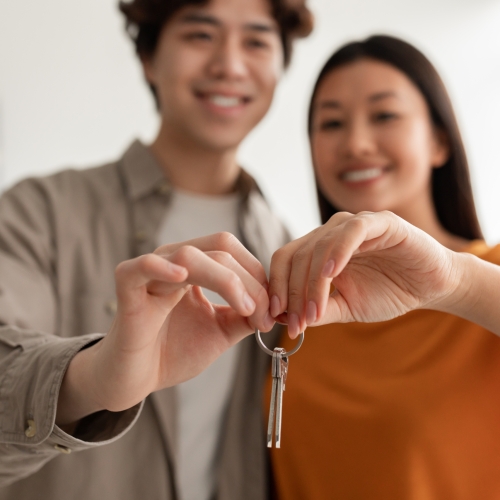 Happy millennial Asian couple showing house key, moving to new apartment together, selective focus. Affectionate young spouses in their house on relocation day. Real estate, rental, mortgage concept